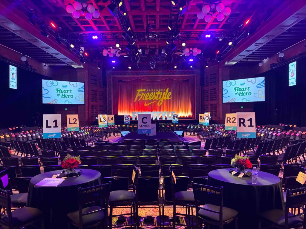 Corporate event ballroom AV production setup with stage lighting, projection screens, and audience seating arranged for a live event.