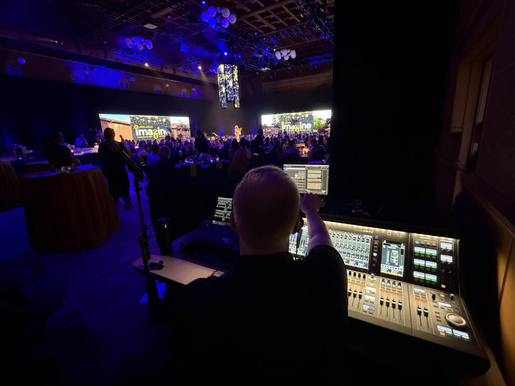 AV show operator running audio and show control during a corporate event in a ballroom with stage screens and seated audience