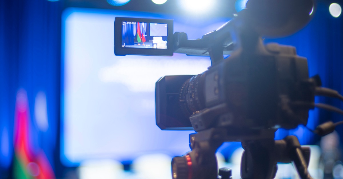 A professional video camera with a screen showing a live event broadcast with blurred stage lighting in the background.