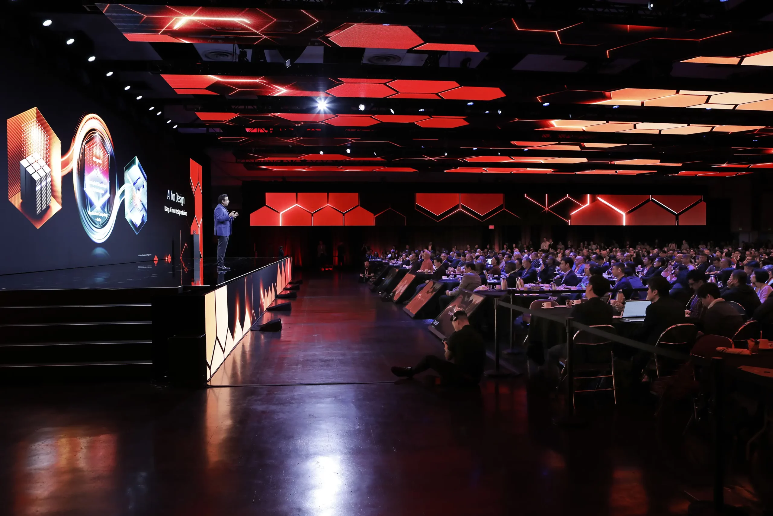 Presenter onstage at CadenceLIVE Silicon Valley with a large audience, LED stage visuals, and red immersive room lighting.