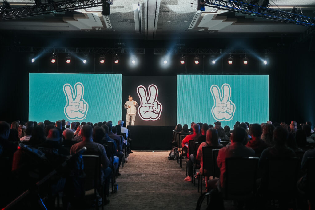 Dutch Bros executive speaking on stage to a packed audience, with a confidence monitor at his feet.