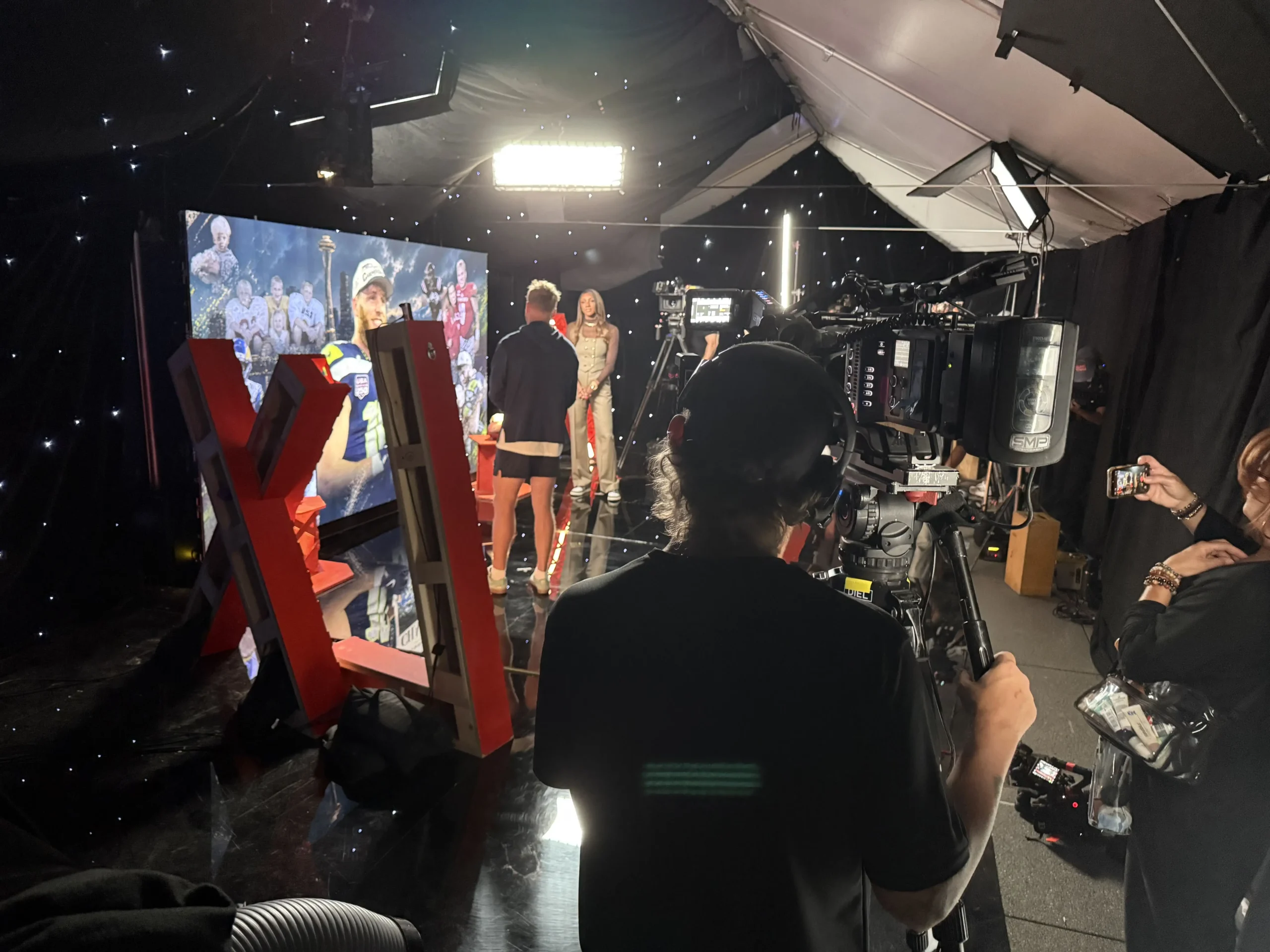 Behind-the-scenes view of the NBC Universal Seattle Seahawks broadcast set with camera crew, LED wall, and star drop drape.